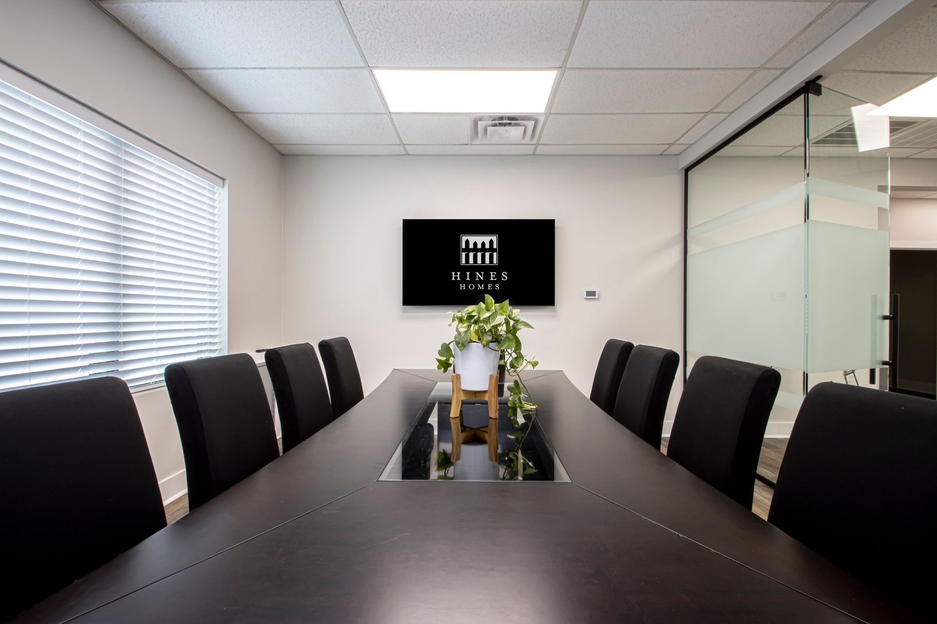 Modern conference room with a long table, black chairs, wall-mounted screen, and flowers in the center