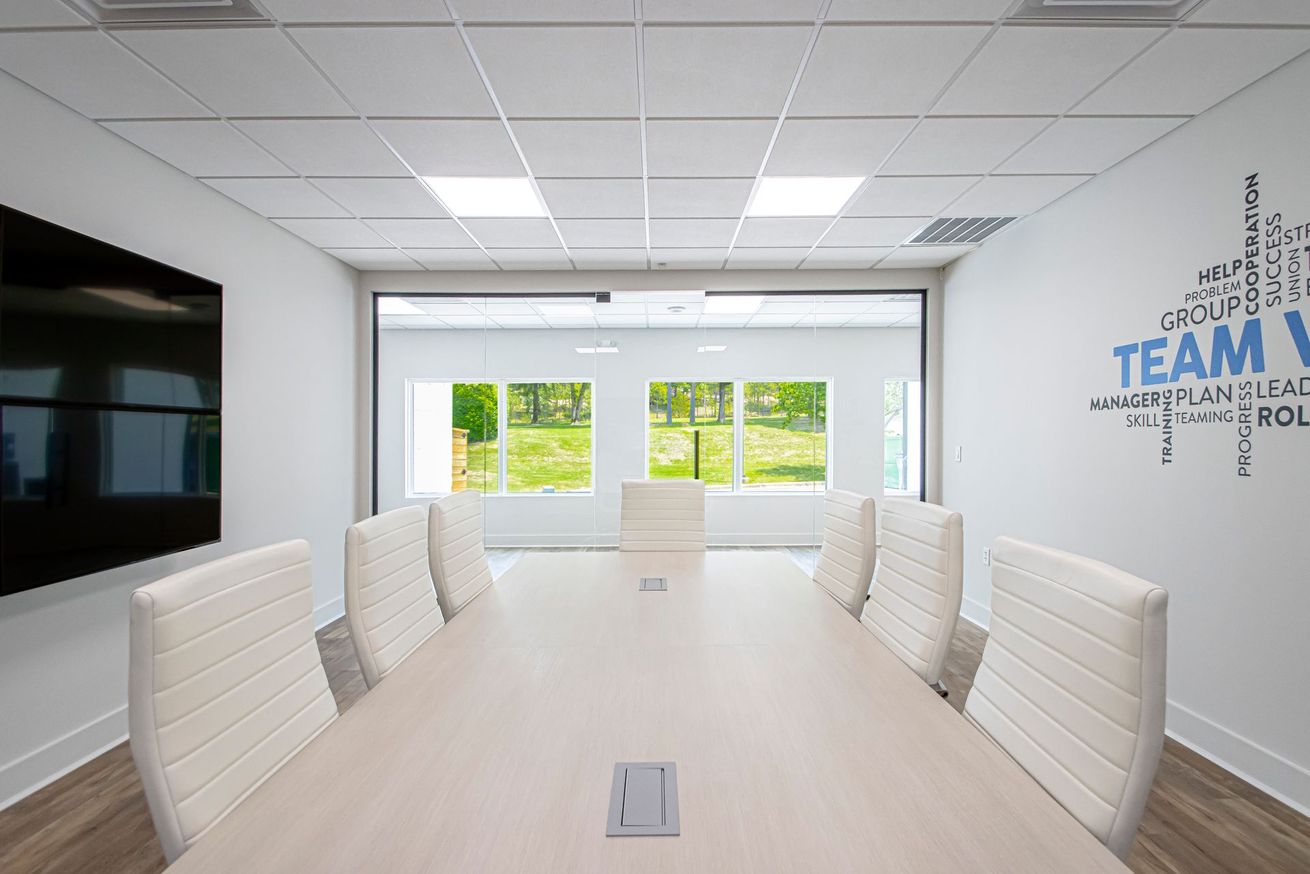 Modern conference room with long table, white chairs, wall-mounted TV, and large windows.