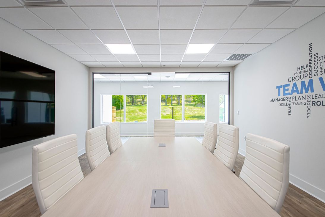 Modern conference room with long table, white chairs, wall-mounted TV, and large windows.