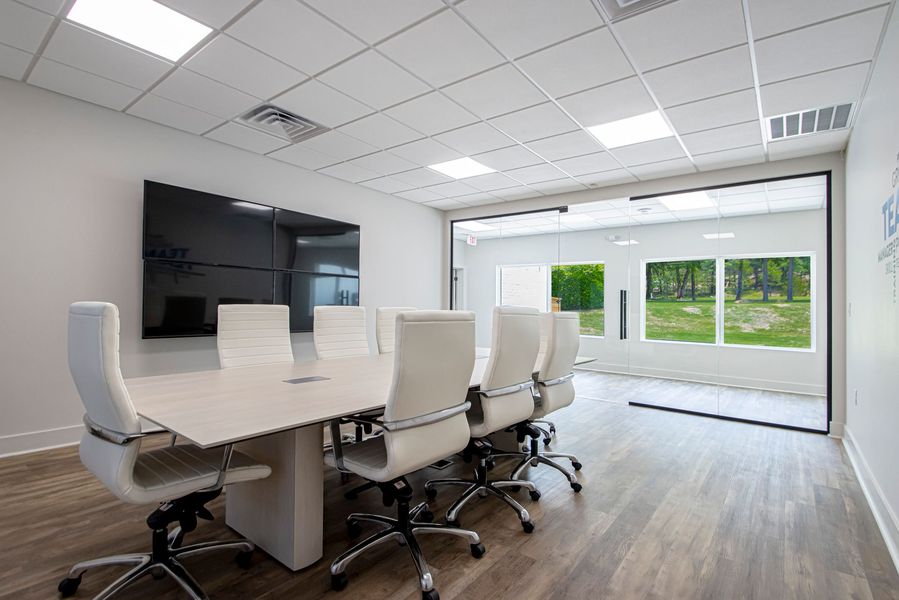 Modern conference room with white chairs, long table, wall display, and large windows overlooking greenery