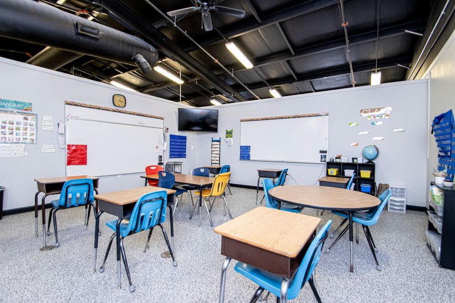 Empty classroom with desks, blue chairs, whiteboards, and a wall-mounted globe under exposed ceiling pipes.