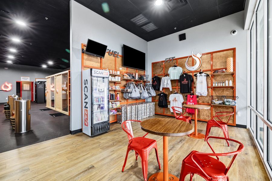 Bright café interior with red chairs, wood tables, shelving, and a drink cooler near the counter.