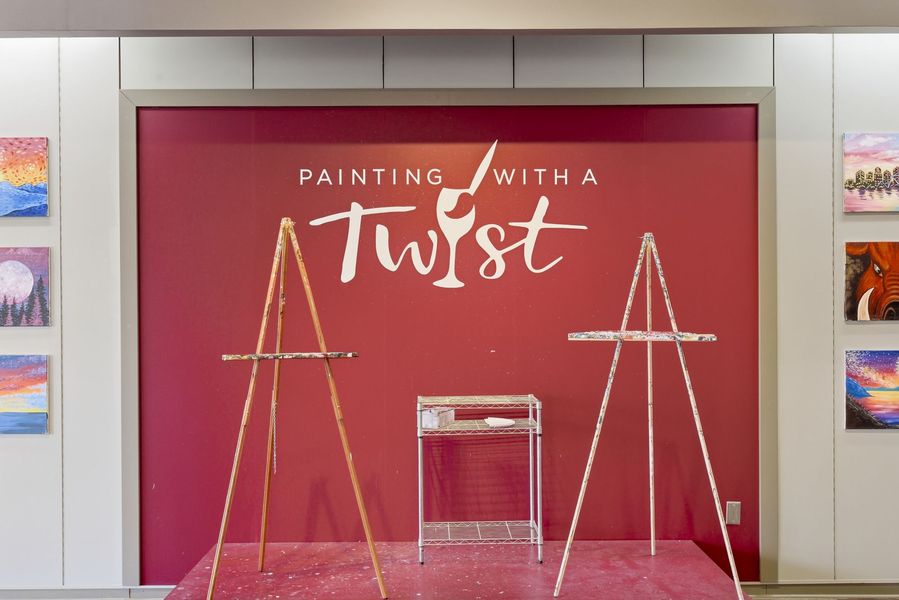 Paint-and-sip display with easels and a podium before a pink backdrop reading “Painting with a Twist”