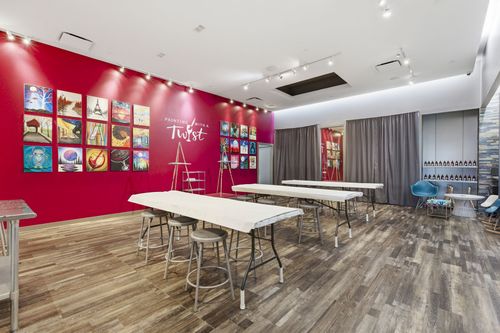 Bright classroom with red accent wall, colorful art, tables, chairs, and gray curtains on the windows.